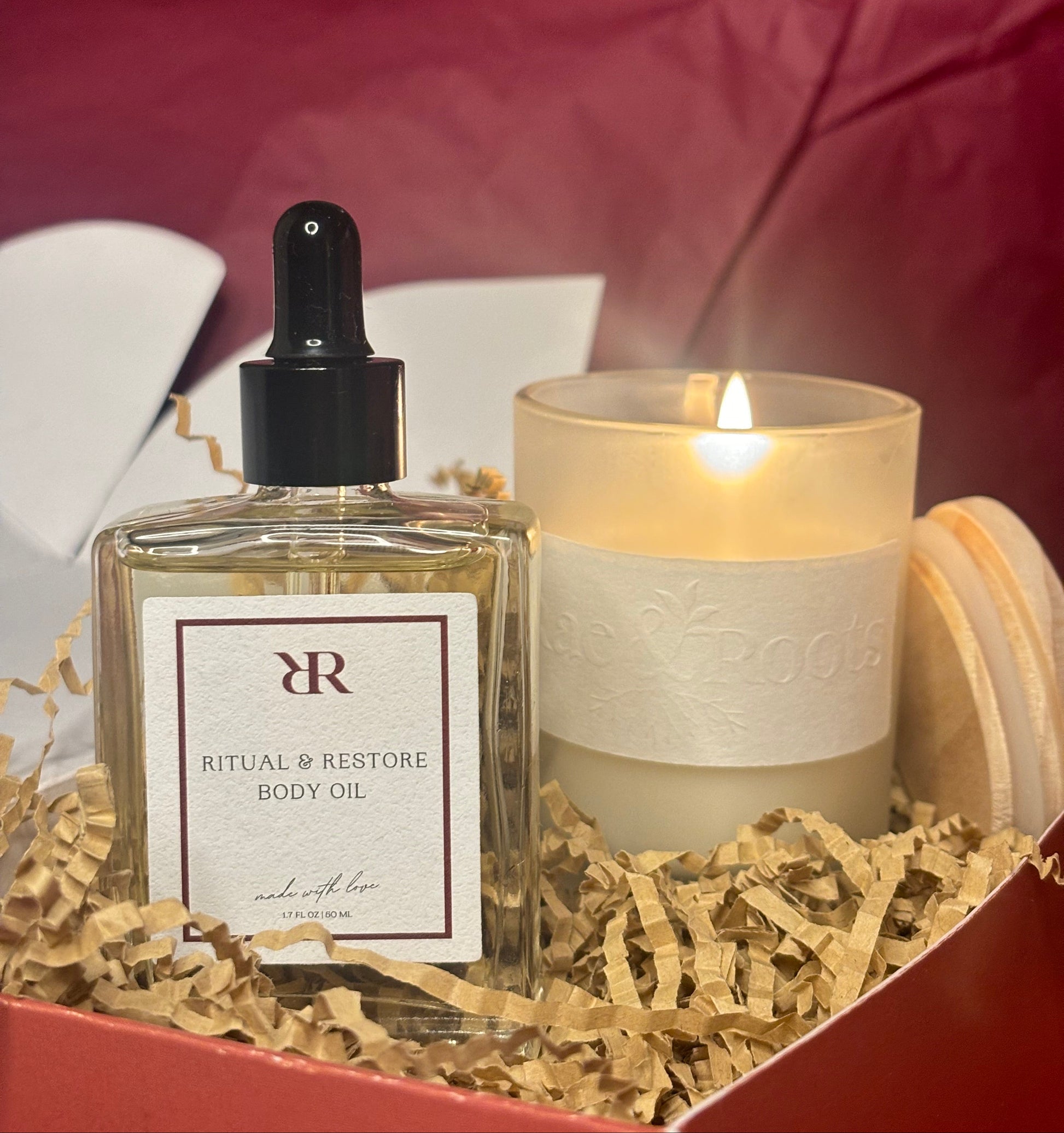 Bottle of Rituals Restore Body Oil with a lit candle in a decorative box on a red fabric background